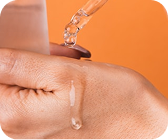 Dropper dispensing clear serum onto the back of a hand against an orange background.