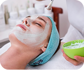Woman lying down with a white facial mask applied and a blue headband, while a person holds a green bowl with cream.