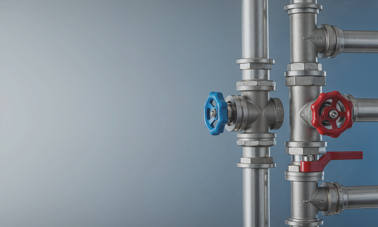 Close-up of industrial metal pipes with red and blue valve handles against a gradient gray-blue background.