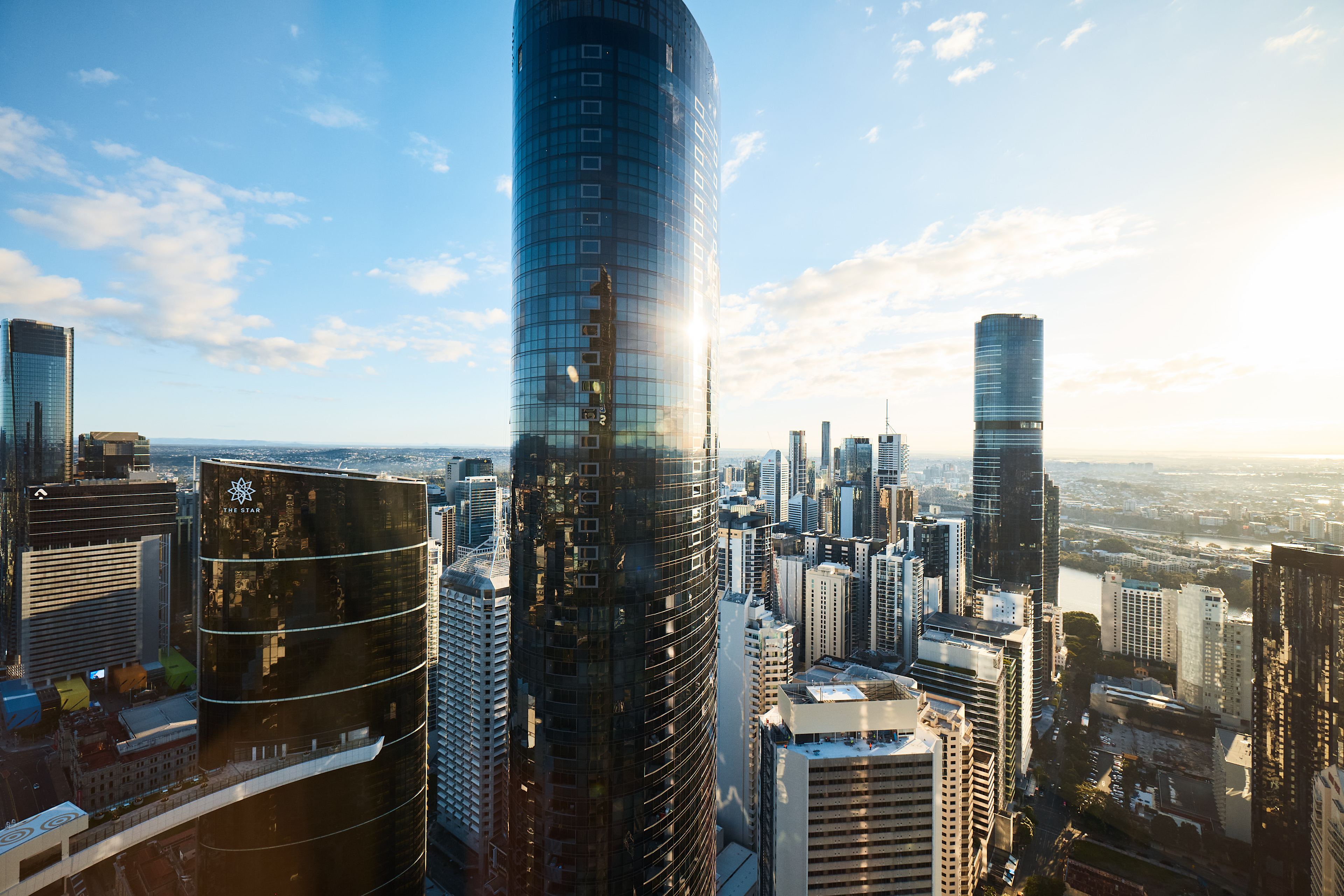 Brisbane city skyline with tall modern skyscrapers, including curved glass towers reflecting sunlight, and the Brisbane River in the background.