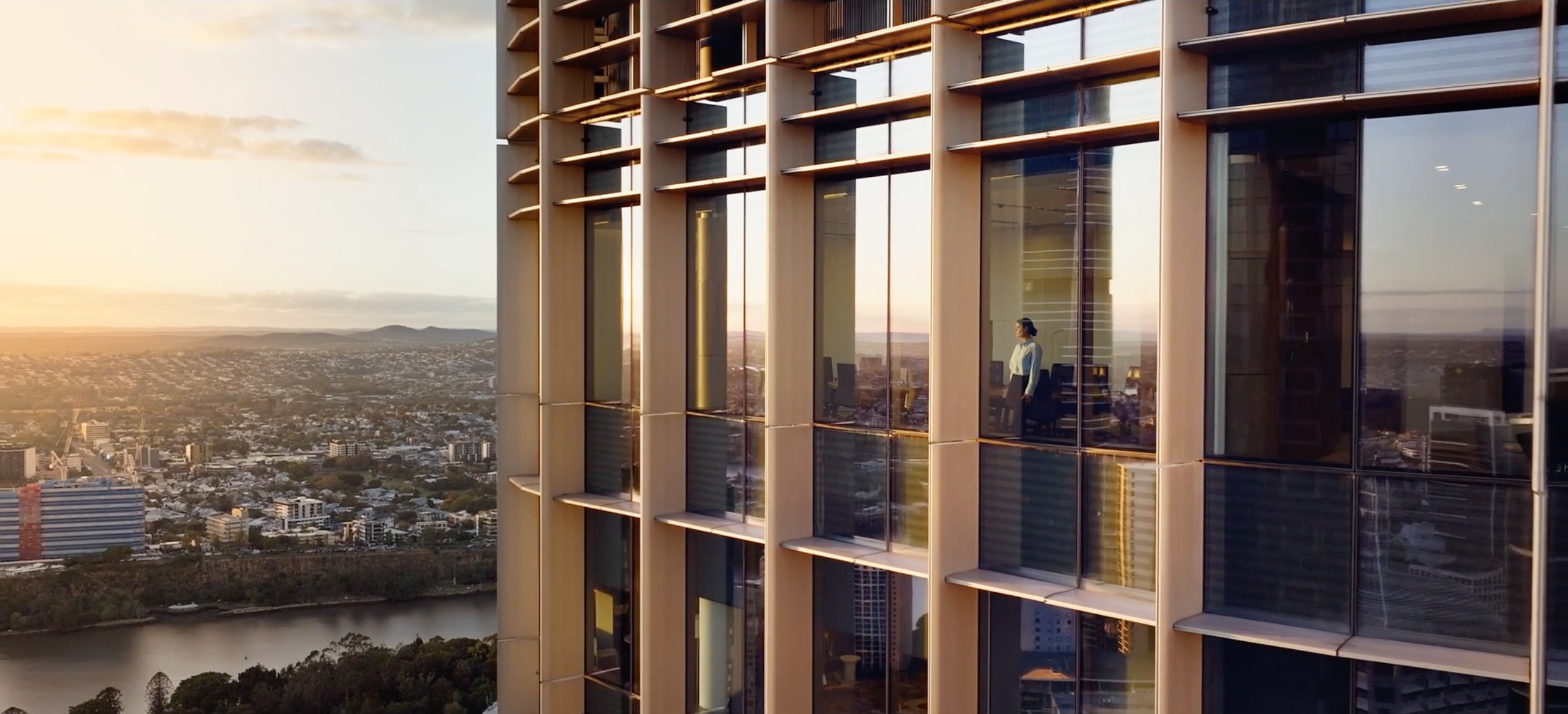 Drone photo taken from outside a  modern high-rise office, with woman looking out large windows over a city skyline.