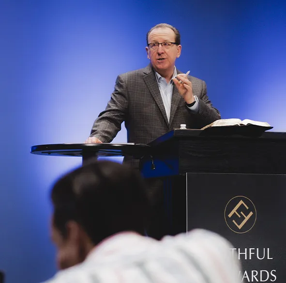 Man in a checkered blazer speaking and gesturing behind a podium with an open book, against a blue background.