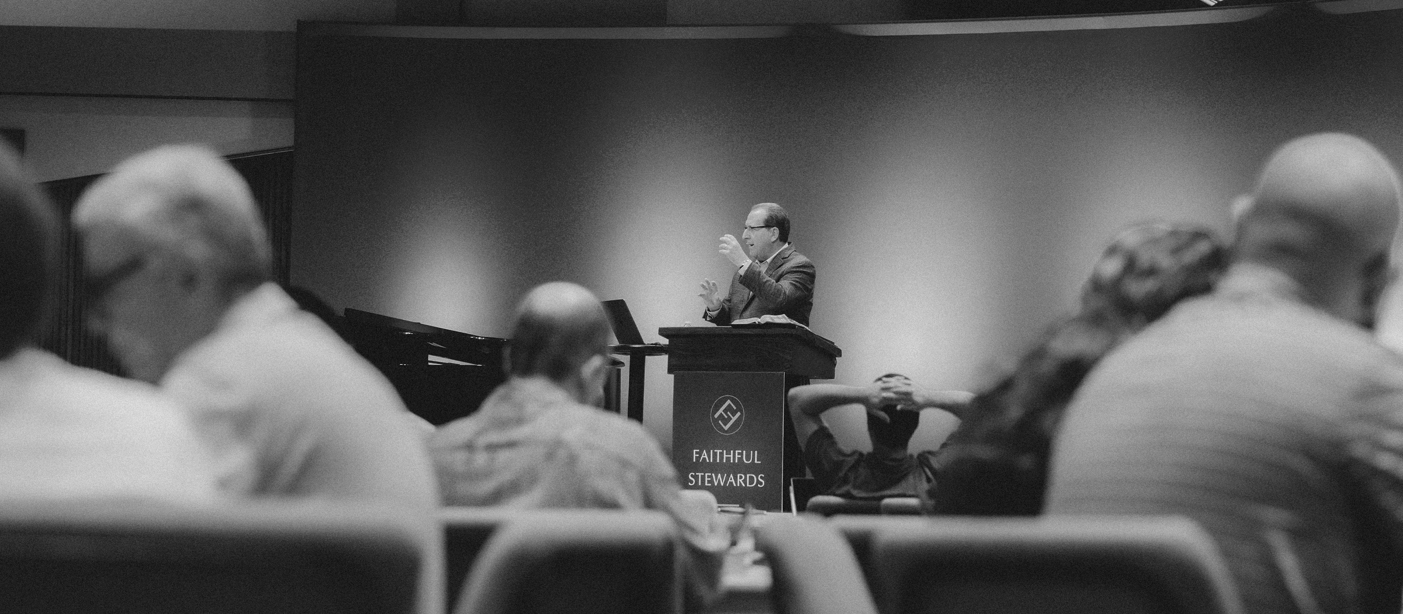 Man speaking at a podium labeled 'Faithful Stewards' in front of an audience in a dimly lit room.