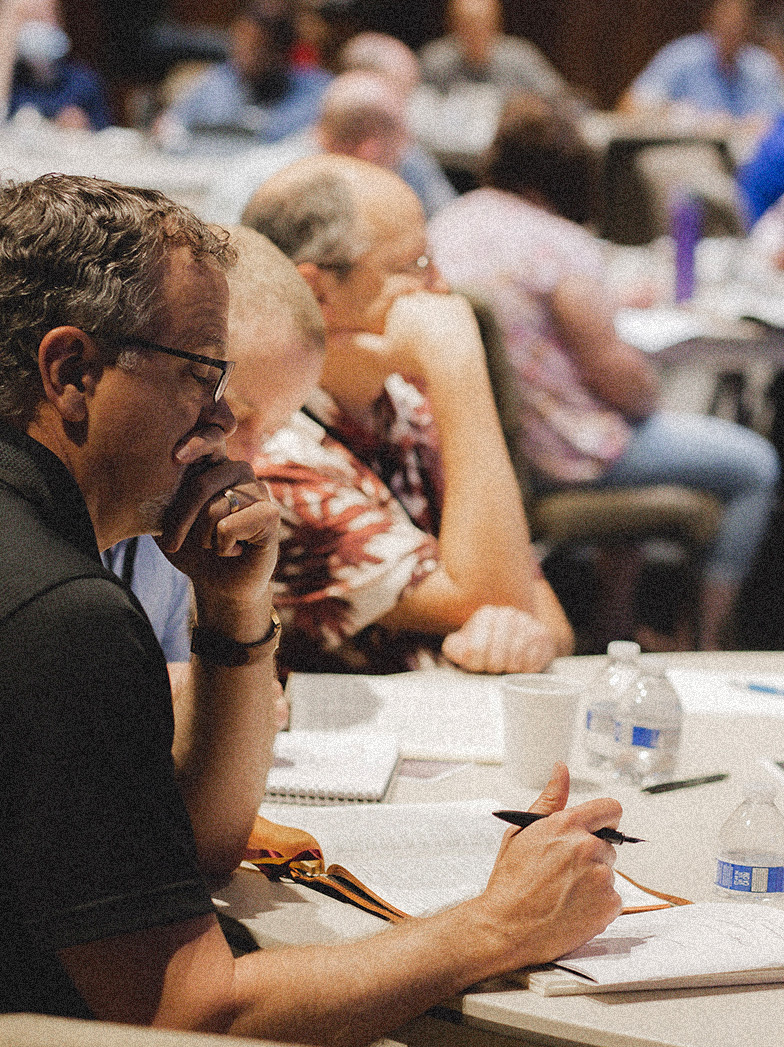Man in glasses writing with a pen at a table during a meeting with other seated participants in the background.