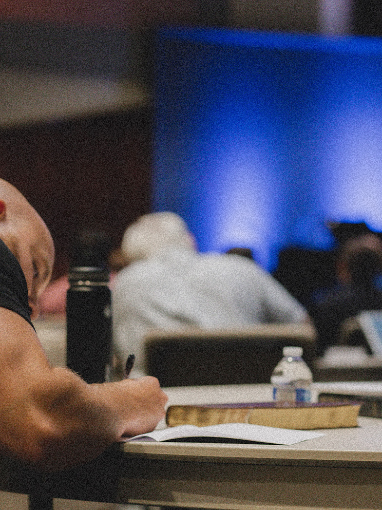 Person writing on paper at a table with a closed book and a water bottle nearby, blurred audience in the background with blue lighting.