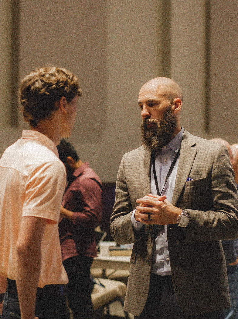 Bearded man in a gray plaid blazer and blue shirt conversing with a young man in a light pink polo shirt in an indoor setting.