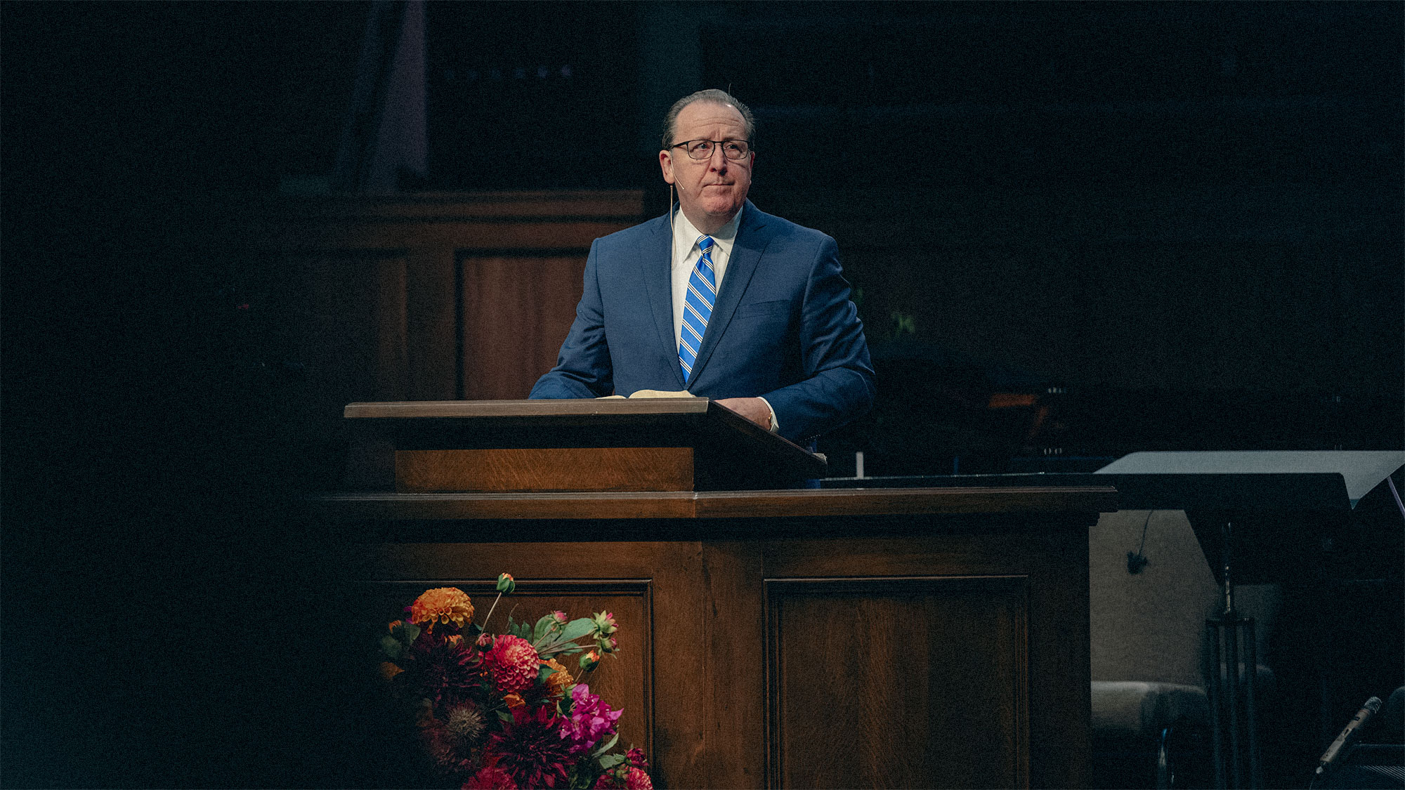 Man in blue suit and striped tie standing behind a wooden podium with flowers in front, speaking in a dimly lit room.