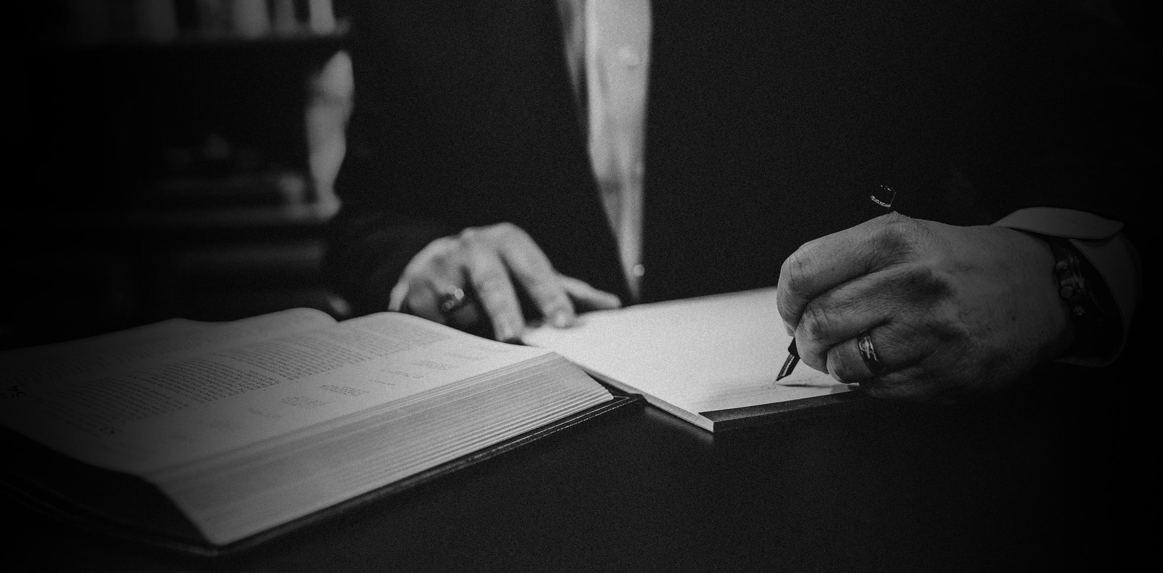 Black and white close-up of a person writing in a notebook with an open book beside them.