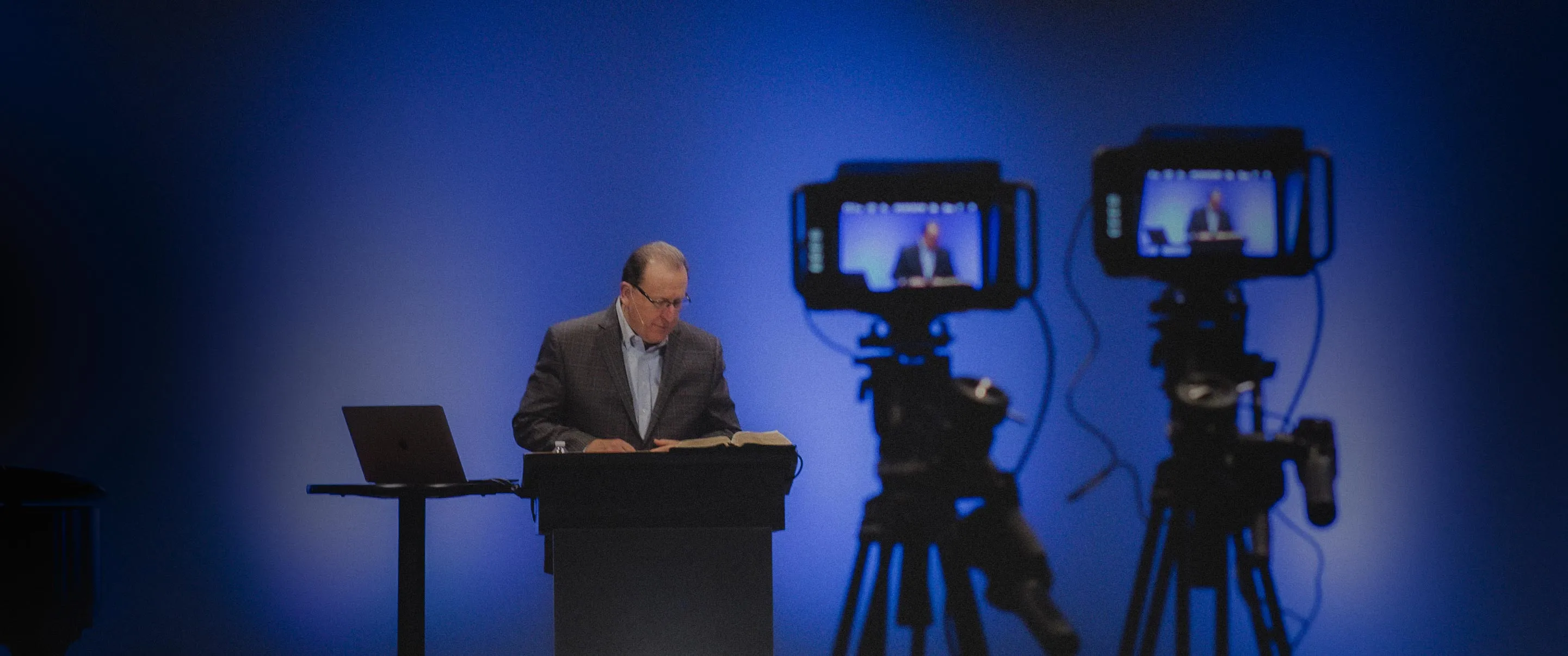 Pastor Pennington standing at a podium reading a book, with a laptop on a side table and two cameras filming him on tripods against a blue background.