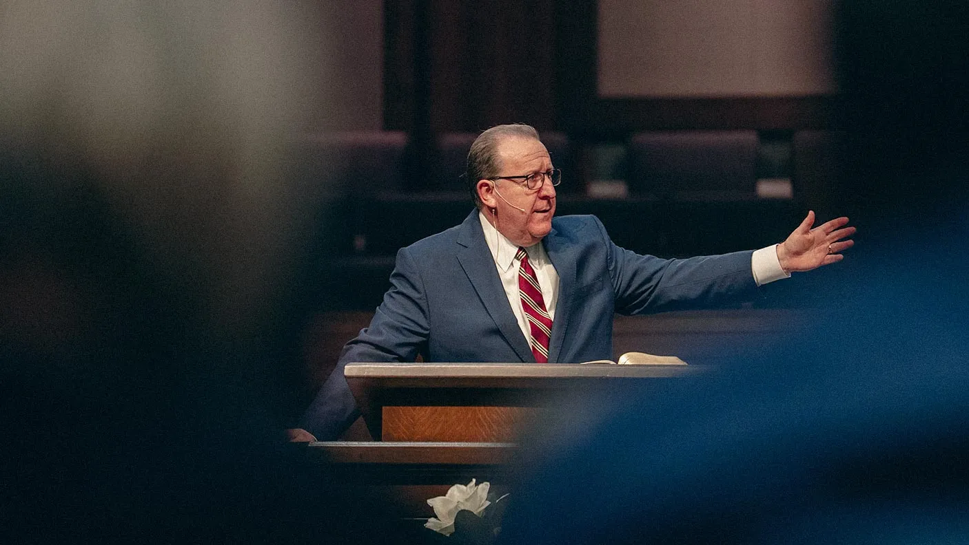 Pastor Pennington in a suit and striped tie speaking at a podium inside a church or auditorium.