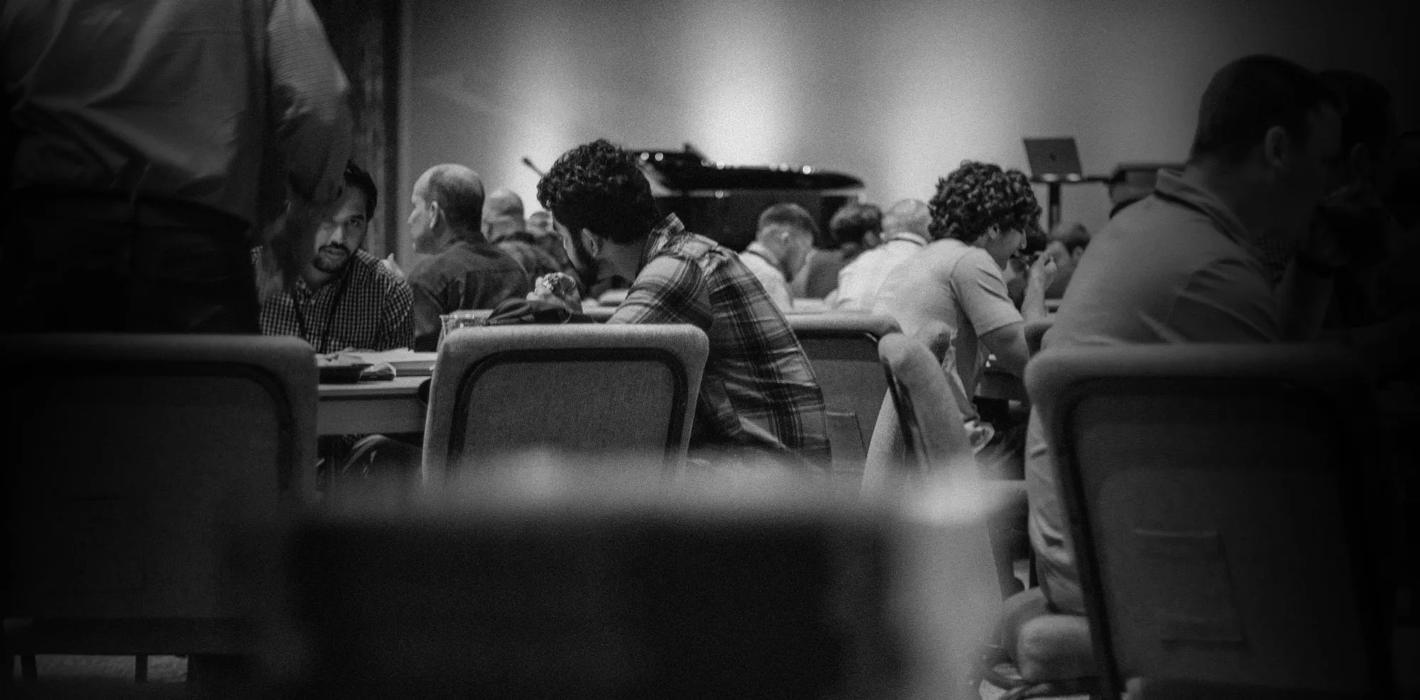 Black and white photo showing people seated at tables in a room, engaged in conversation and eating.