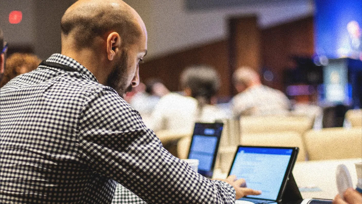 Man in checkered shirt working on a tablet in a conference room with other attendees seated in the background.