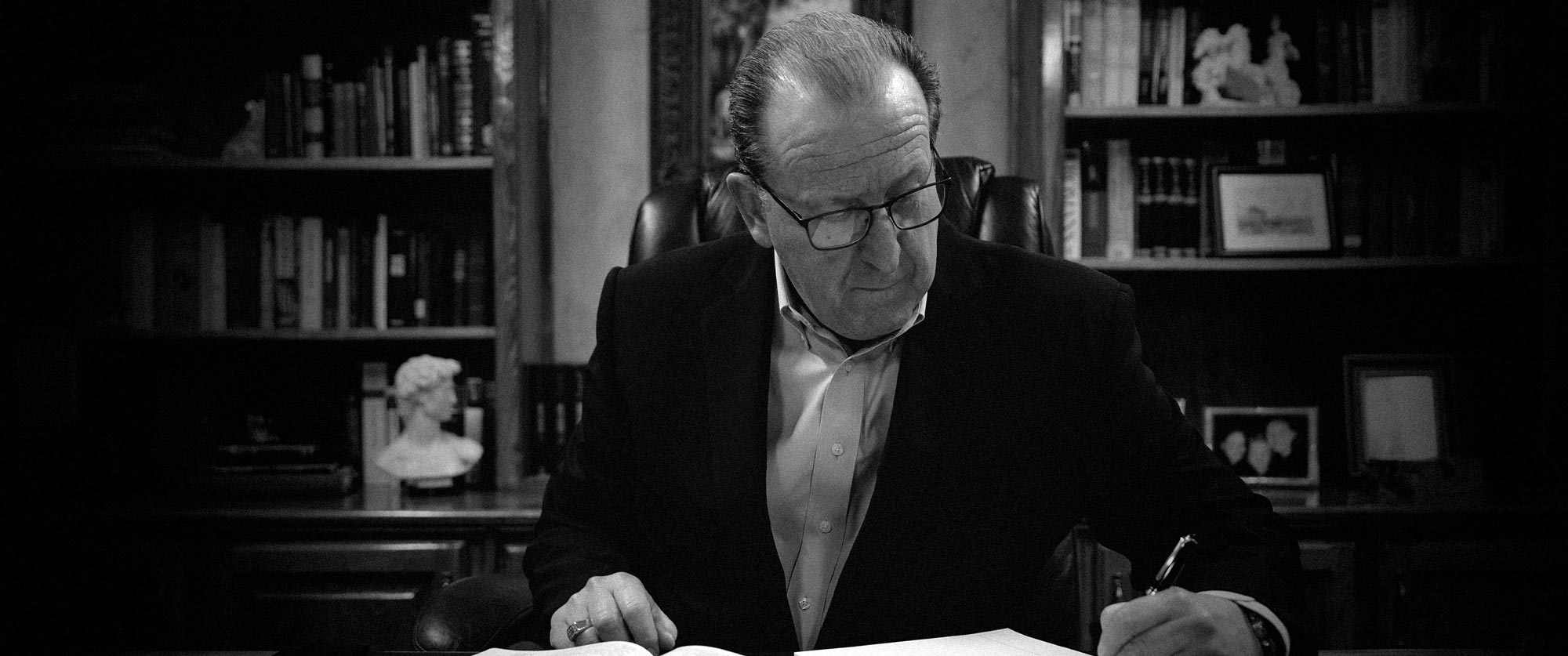 Pastor Pennington wearing glasses and a suit jacket sitting at a desk, writing on papers with bookshelves in the background.