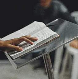 Close-up of a hand turning pages of an open Bible on a transparent lectern.