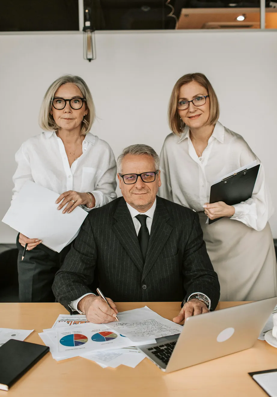 Three business professionals, two women standing and a man sitting at a desk with documents, charts, and a laptop.