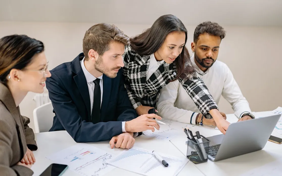Four business professionals collaborating and looking at a laptop screen surrounded by charts and documents on a white table.