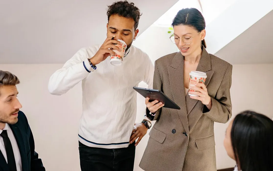 Four colleagues in a casual office setting, two standing with coffee cups and a tablet, two seated and engaged in discussion.