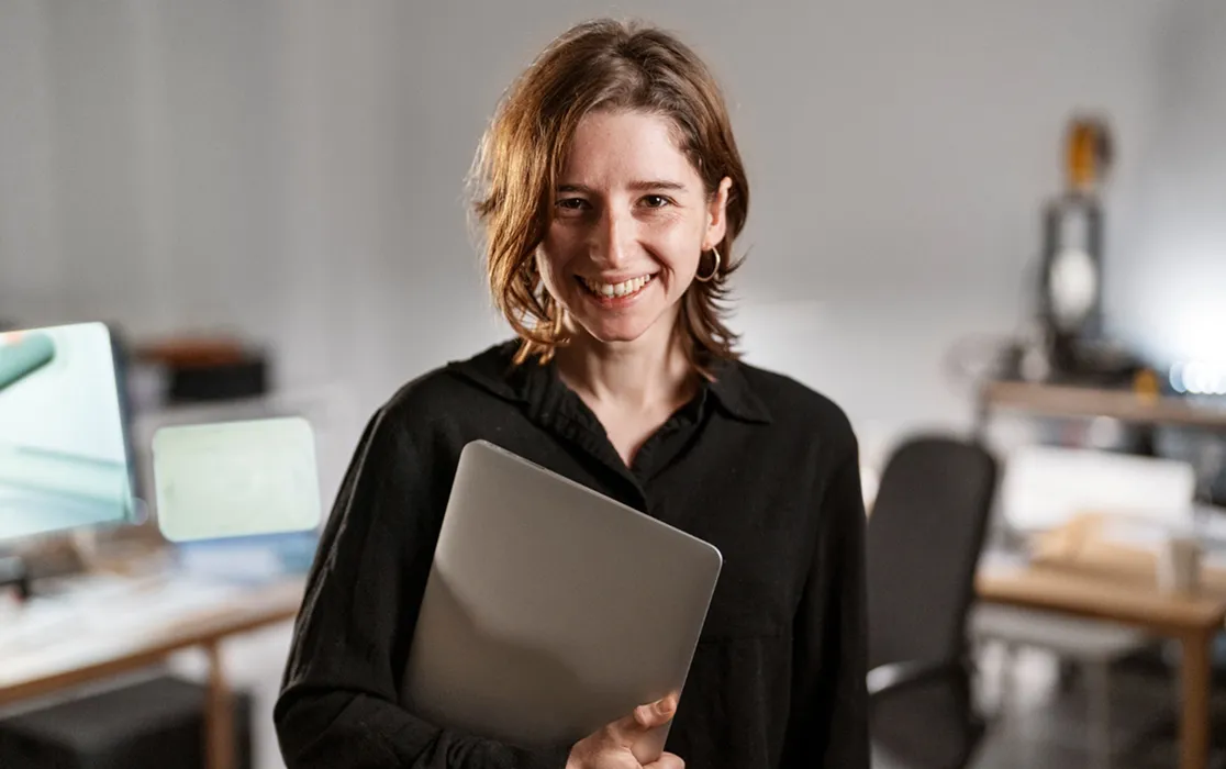 Smiling woman in a black shirt holding a closed laptop in an office setting.