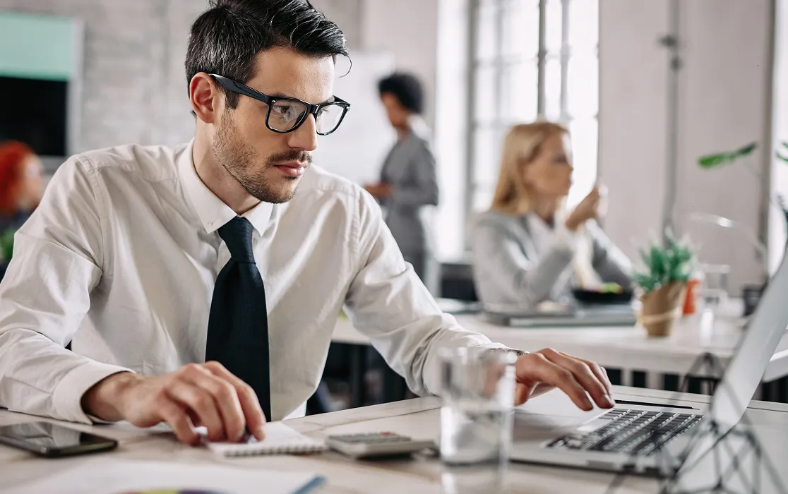 Young man in glasses and white shirt working on a laptop at a desk in an office.