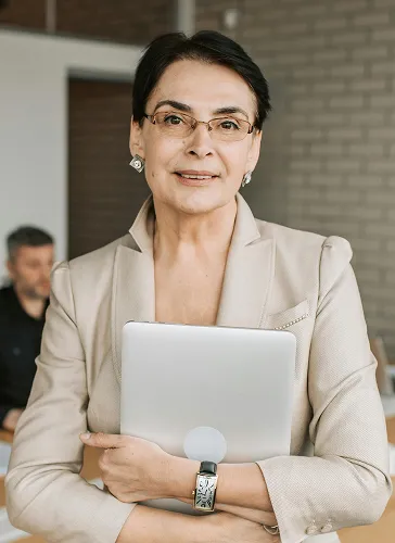 Professional woman in glasses and beige blazer holding a laptop with folded arms, smiling at the camera.