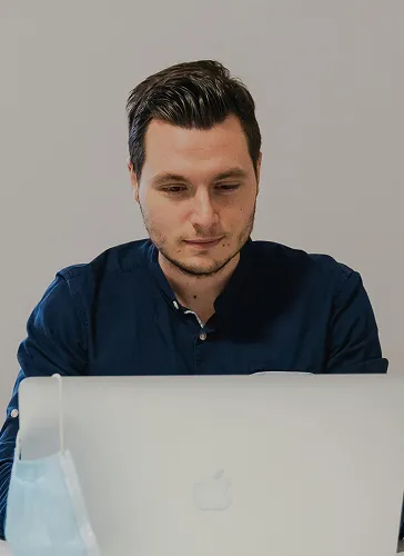 Man with dark hair focused on a laptop screen, wearing a dark blue shirt with a face mask hanging on the side.