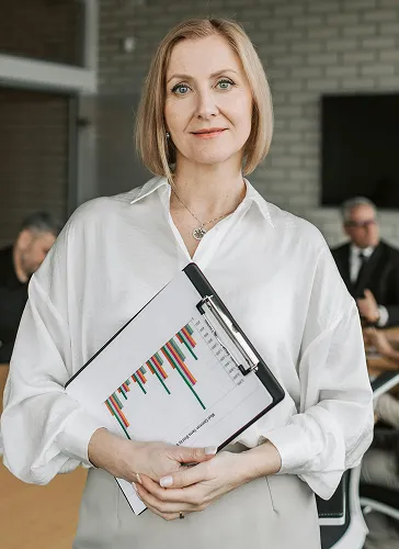 Professional woman in a white blouse holding a clipboard with a bar chart, with blurred office colleagues in the background.
