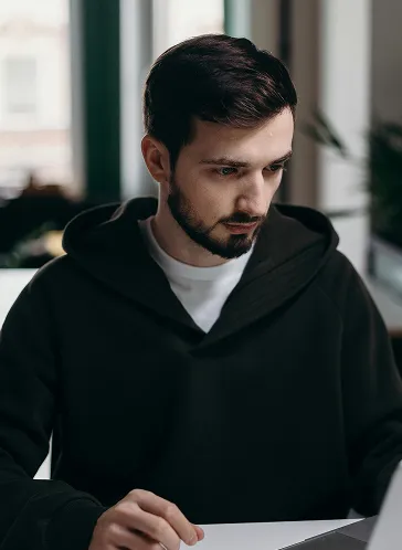 Young man with dark hair and beard wearing a black hoodie, looking down at a laptop in a softly lit room.