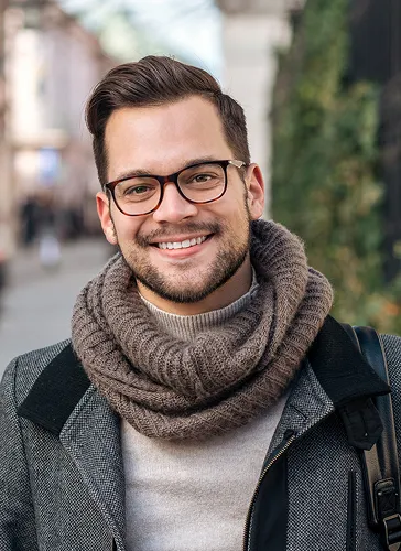 Smiling man with glasses wearing a brown knitted scarf and gray coat outdoors.