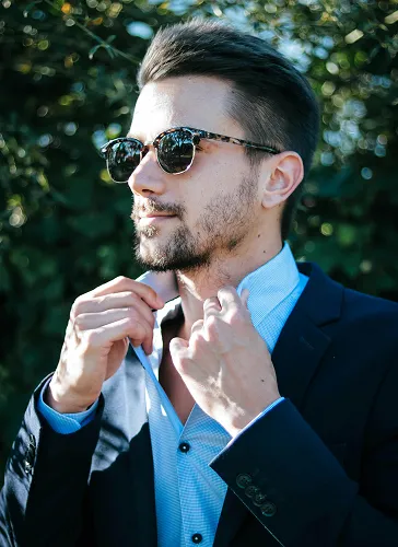 Man wearing sunglasses adjusting the collar of his light blue shirt under a dark blazer outdoors with greenery in the background.