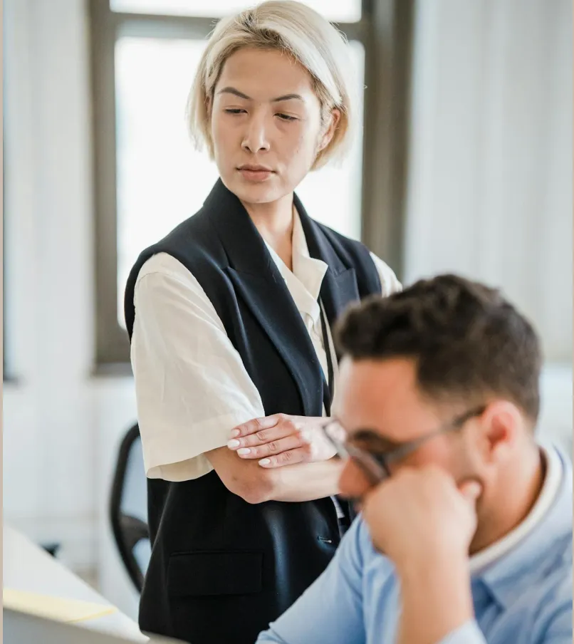 Woman with short blonde hair and black vest stands with arms crossed looking at a man wearing glasses who is sitting and resting his chin on his hand in an office.