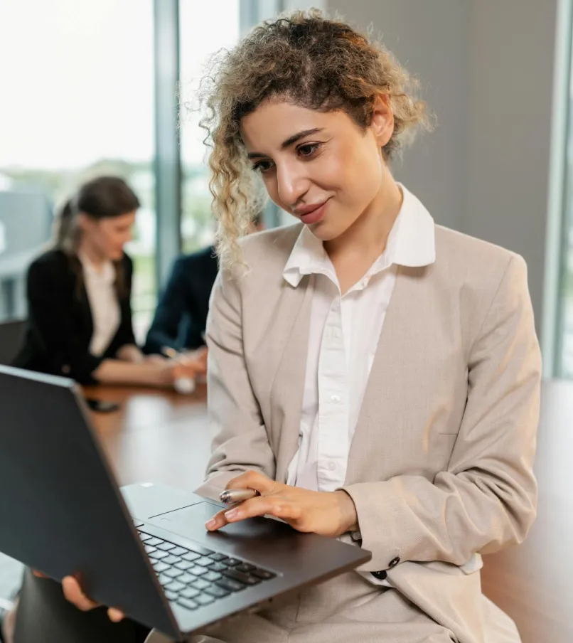 Businesswoman in a beige suit using a laptop with two blurred colleagues in the background.