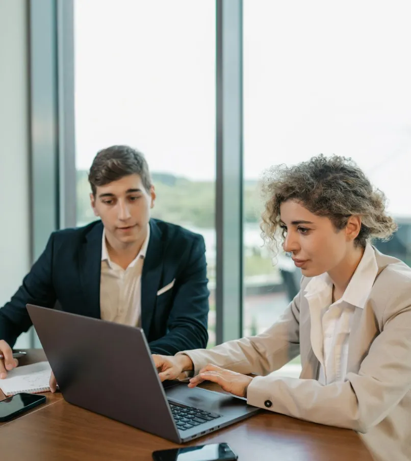 Two business professionals, a man and a woman, working together on a laptop in a modern office with large windows.