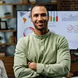 Smiling man in a green hoodie standing with arms crossed in an office with charts on a screen behind him.