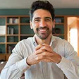 Smiling man with dark hair and beard sitting indoors with hands clasped.