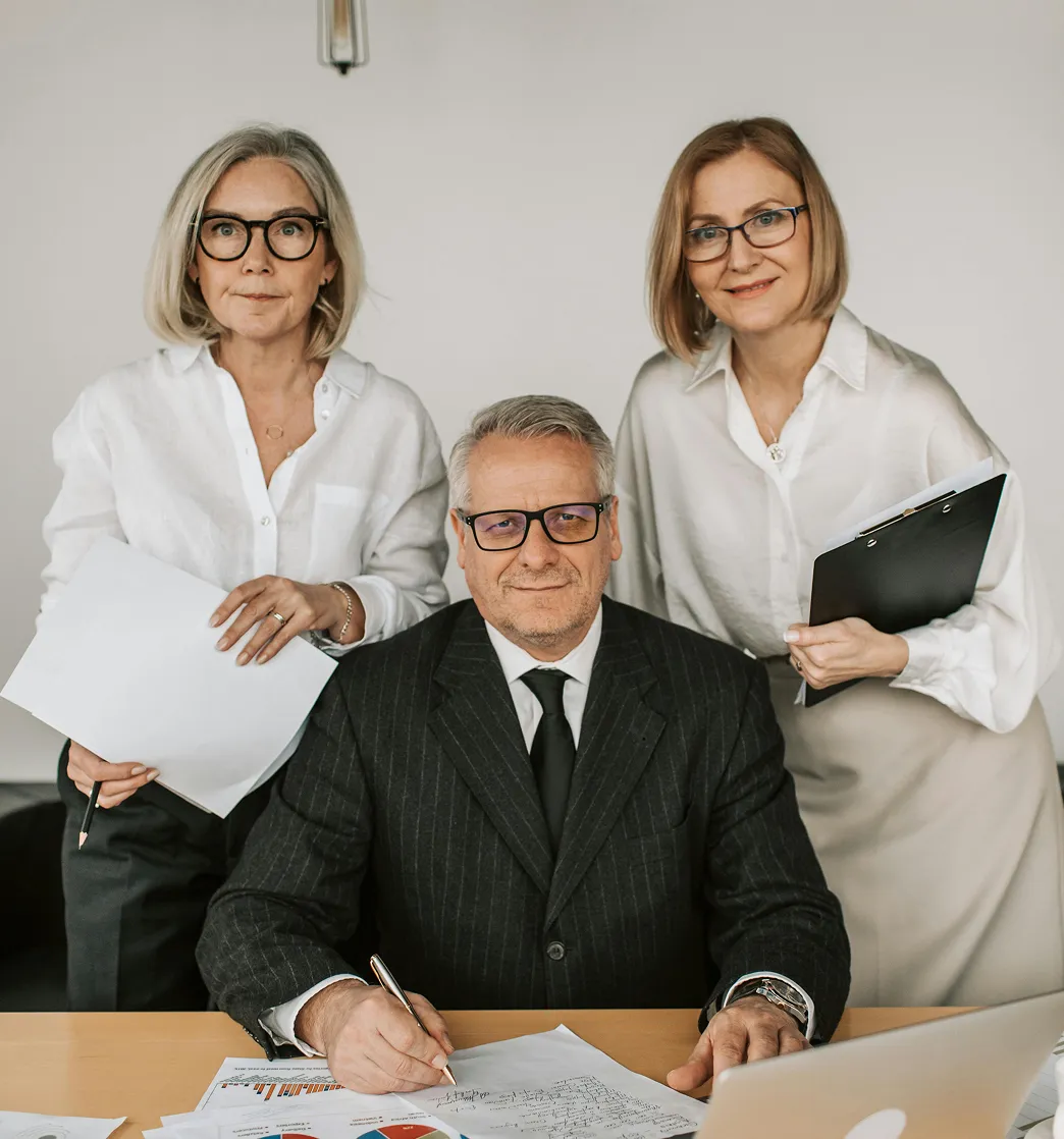 Three business professionals with glasses, two women standing and one man seated at a desk with documents and a laptop.