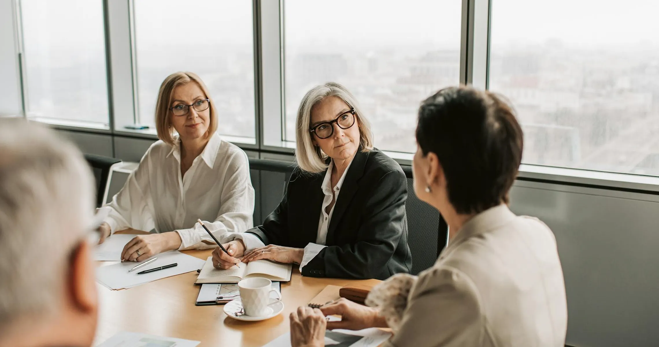 Four professionals engaged in a meeting around a table in a bright office with large windows overlooking a cityscape.