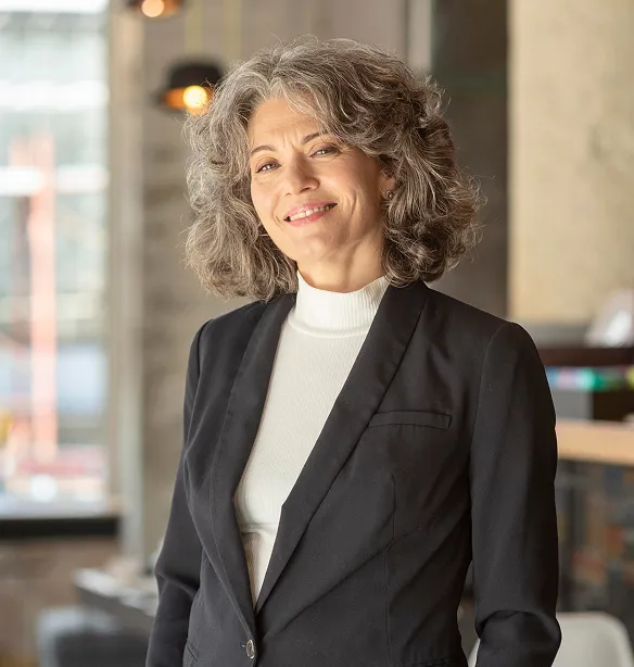 Smiling middle-aged woman with curly gray hair wearing a black blazer and white turtleneck in a modern office.