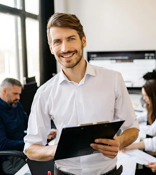 Smiling man in white shirt holding a clipboard in an office with colleagues working in the background.