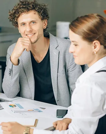 Man in a gray blazer smiling and sitting at a table with a woman writing on a clipboard.