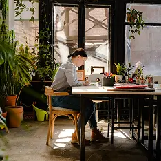 Person sitting at a desk inside a sunlit room filled with plants, focused on working with a laptop and papers.