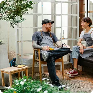 Two baristas in aprons sitting and chatting in a coffee shop with plants and large windows.