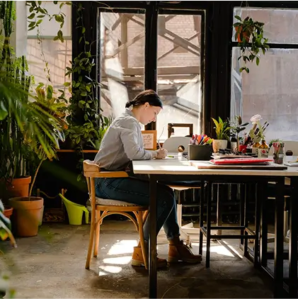 Person sitting at a bright table near large windows, writing with plants surrounding the workspace.
