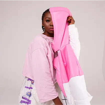 Woman with braided hair wearing a light pink cropped sweatshirt and patterned pants, holding a pink and white jacket partially covering her face.