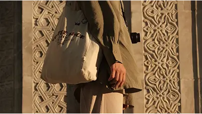 Person wearing an olive green shirt and beige pants holding a camera and a white tote bag with text, standing in front of a carved stone wall.