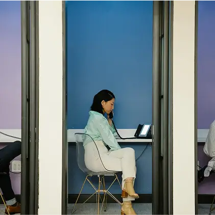 Woman sitting on a transparent chair in a small blue-walled office booth, speaking on a phone.