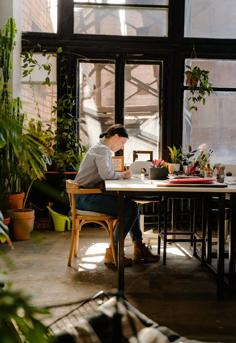 Woman sitting at a table writing with plants surrounding large sunlit windows in a cozy indoor workspace.