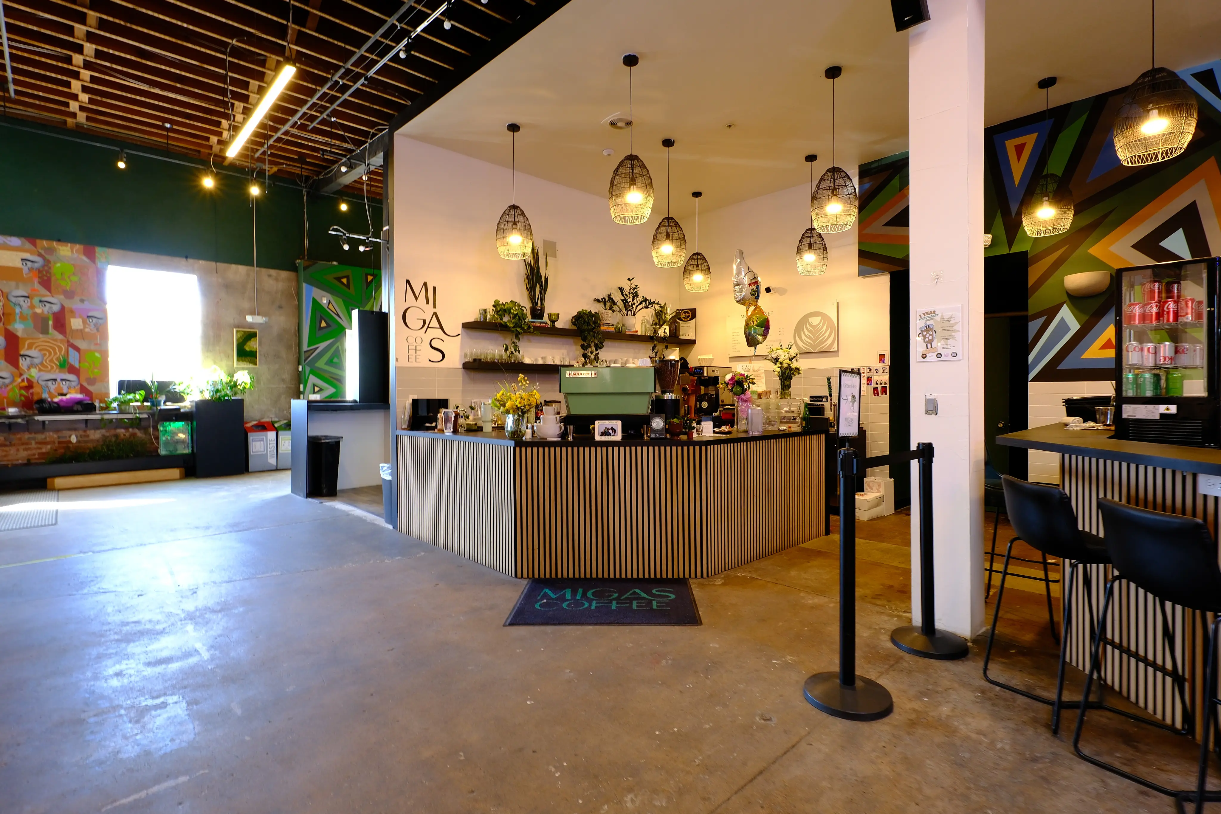 Interior of a modern coffee shop with wooden counter, hanging lights, plants, and colorful geometric wall art.