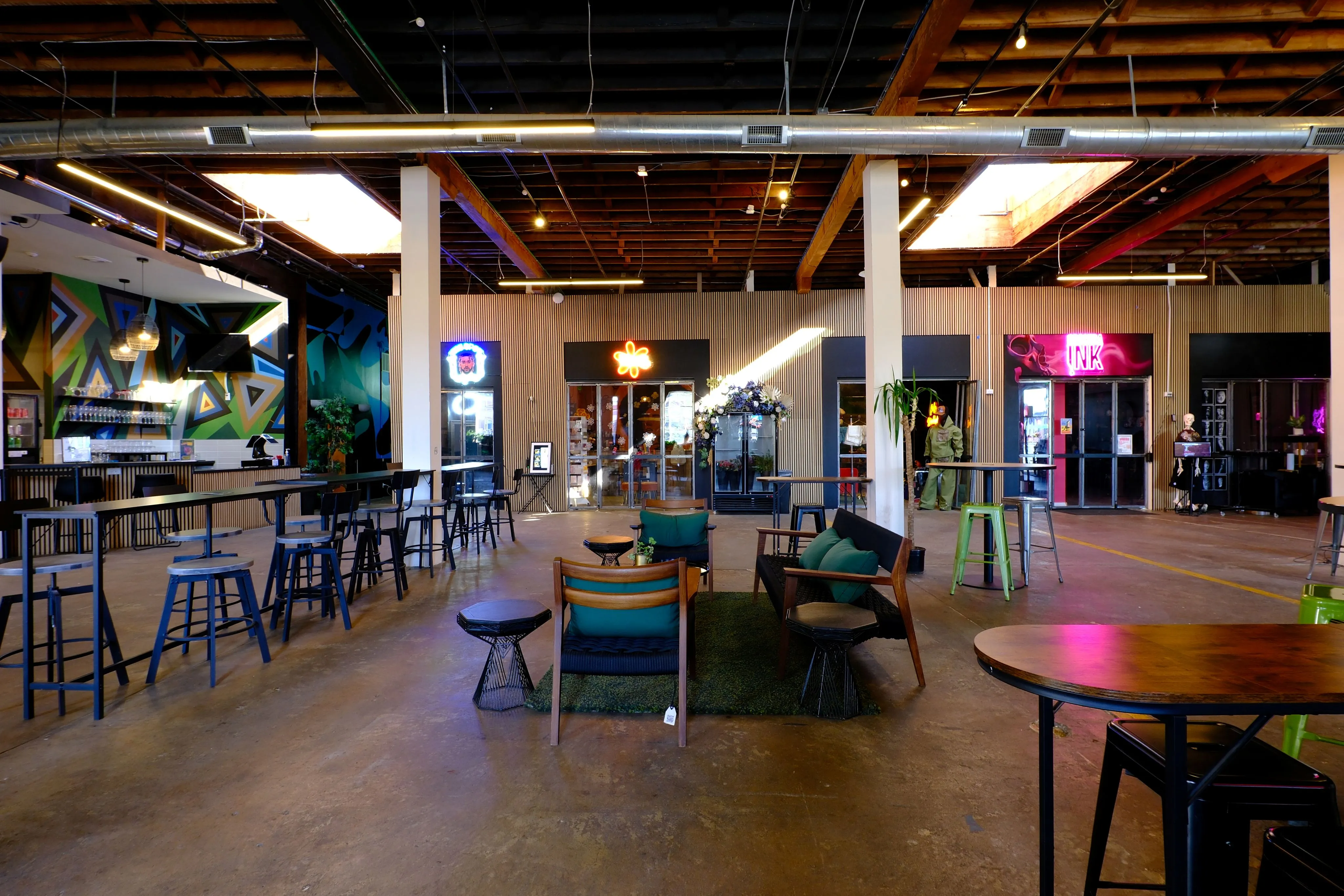 Spacious industrial-style café with wooden ceiling beams, neon signs, high tables with stools, and a cozy seating area with chairs and cushions on a green rug.