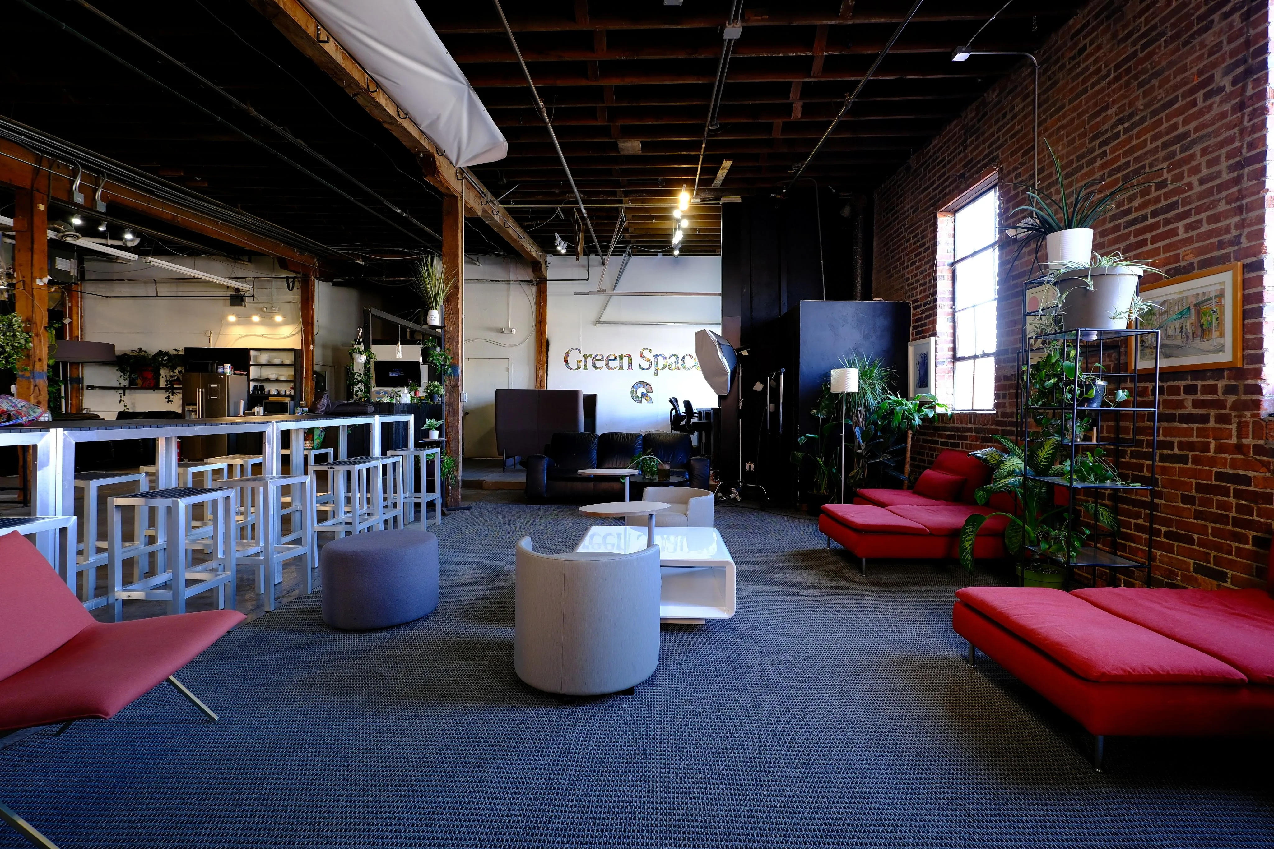 Modern open office lounge with red sofas, gray chairs, high stools at a bar, and green plants against exposed brick walls.