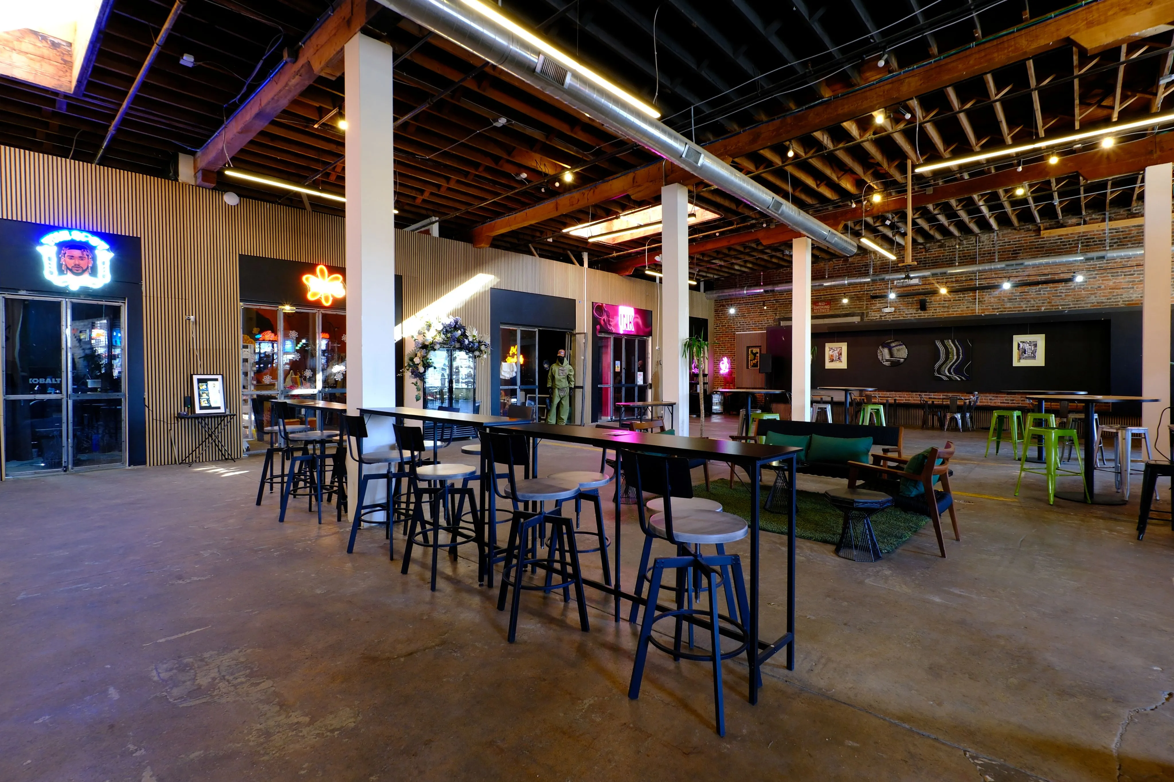 Spacious modern industrial interior with high wooden ceiling, long black tables with stools, and neon signs on walls.